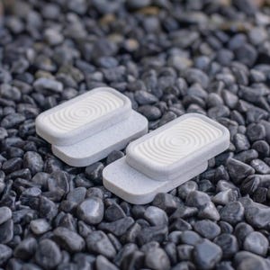 Puede incluir: Dos objetos rectangulares de color gris claro con textura moteada, cada uno con un panel central blanco elevado con un patrón de círculos concéntricos. Los objetos tienen un diseño en capas con una base más ancha y una sección superior ligeramente más pequeña.