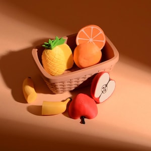 Könnte beinhalten: Ein kleiner brauner Kunststoffkorb mit Miniatur-Radiergummis in Obstform, darunter eine gelbe Ananas, eine Orangenscheibe und eine runde Orange. Außerhalb des Korbes liegen zwei gelbe Bananenstücke und zwei rote Apfelstücke.