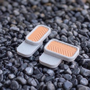 May include: Two rectangular objects featuring a light gray speckled base and a raised peach-colored insert with a chevron pattern. Each piece has a smooth, rounded edge design, with one object positioned slightly behind and to the right of the other.