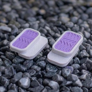 Puede incluir: Dos objetos rectangulares con una base de color blanco roto moteado y una superficie superior elevada de color morado vibrante con un patrón en relieve de ondas repetidas. Los artículos tienen un acabado mate y bordes redondeados.