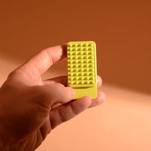 Könnte beinhalten: Ein rechteckiges gelbes Objekt mit einer strukturierten Oberfläche, das im oberen Bereich ein Gitter aus kleinen, erhabenen, pyramidenförmigen Noppen und im unteren Bereich eine glatte, flache Oberfläche aufweist, konzipiert für taktile Zwecke.