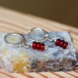 May include: A pair of silver hoop earrings with a dangling cluster of red beads. The earrings are displayed on a multi-colored stone with a wooden background. The earrings are a simple yet elegant accessory.
