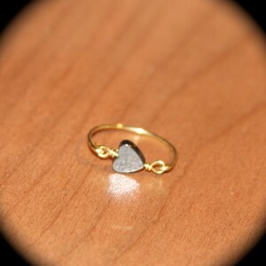 May include: A delicate gold-colored ring featuring a small, heart-shaped, gray stone. The heart is secured with wire, and the ring band is thin and smooth. The ring is set against a warm, brown background.