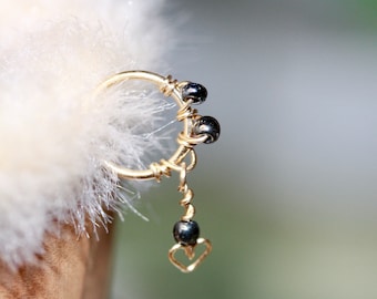May include: A gold hoop earring with a dangling charm. The charm features a heart shape and three black beads.