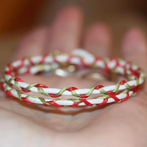 May include: A white bracelet with red and green woven accents. The bracelet is made of two strands, each with a white base and a pattern of red and green threads woven around it. The bracelet is held in a hand.