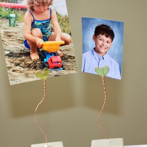 May include: Two photo holders with copper wire stems and heart-shaped green accents. Each holds a photograph. One shows a child playing with a toy truck, the other a portrait of a smiling boy. The bases are white.