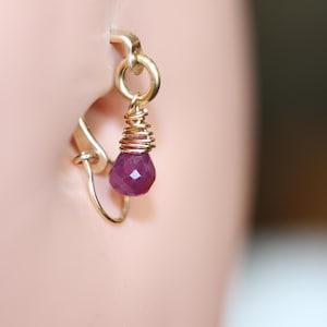 May include: A gold-colored earring with a small, faceted, deep pink gemstone. The gemstone is suspended from a coiled wire and a small hoop that attaches to the ear. The earring is shown in a close-up view.