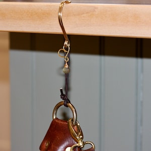 May include: A brass-colored hook with a decorative swirl design, a small green bead, and a leather strap. The hook is attached to a brown leather item with a gold-colored clasp.
