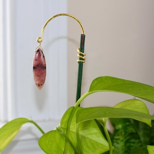 May include: A decorative plant stake featuring a pink and black gemstone pendant suspended from a gold-colored metal arch. The stake is green with a gold spiral accent. Lush green leaves of a houseplant are in the foreground.