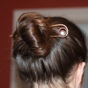 May include: A woman's brown hair is styled in a bun and held in place with a copper hair pin. The pin has a spiral design with a small, round, white accent.
