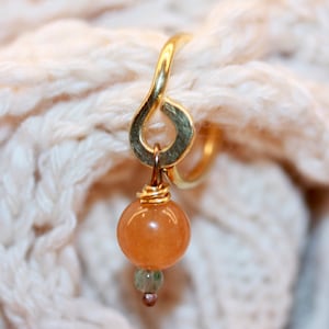 May include: A gold metal ring with a teardrop-shaped charm. The charm is attached to a small, round, orange-colored stone.