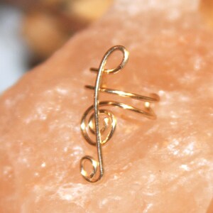 May include: Gold treble clef ear cuff. The cuff is made of wire and is shaped like a treble clef. It is a delicate and stylish piece of jewelry.