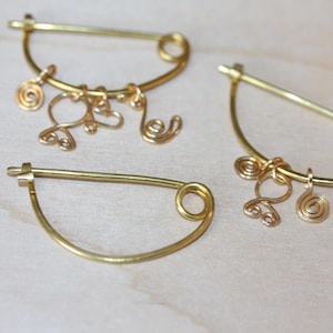 May include: Three gold-colored safety pins with decorative wire charms. The pins are arranged on a light-colored wooden surface. The charms include spiral and abstract shapes, adding a touch of elegance to the jewelry.