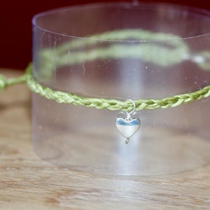 May include: A green braided bracelet with a silver heart charm hanging from it. The bracelet is displayed on a clear glass surface.