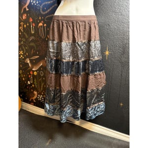 May include: A tiered midi skirt featuring alternating horizontal bands of brown fabric and shimmering dark gray velvet. The skirt has an elasticized waistband and a flared silhouette that falls below the knees, constructed with multiple stitched panels.