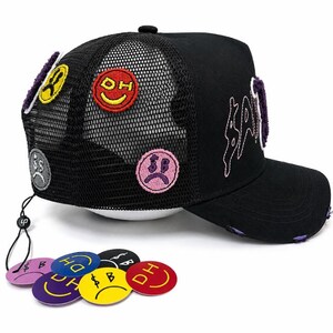 May include: A black trucker hat with a mesh back, featuring embroidered patches of colorful smiley faces and sad faces. The front displays purple stitched lettering. A string attached to the back holds several circular, colorful plastic tags.