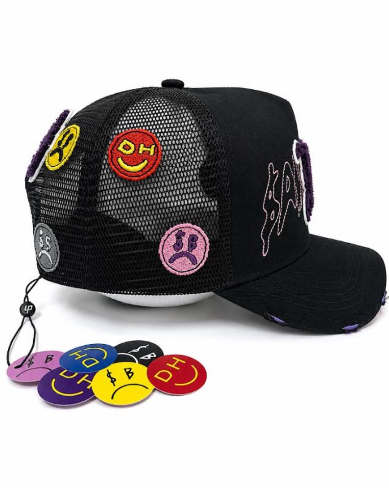 May include: A black trucker hat with a mesh back, featuring embroidered patches of colorful smiley faces and sad faces. The front displays purple stitched lettering. A string attached to the back holds several circular, colorful plastic tags.