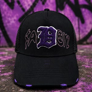 May include: A black trucker hat featuring a mesh back and a distressed brim with purple accents. The front panel displays a large, textured purple letter B patch with white trim, flanked by embroidered text that includes a sad face icon.