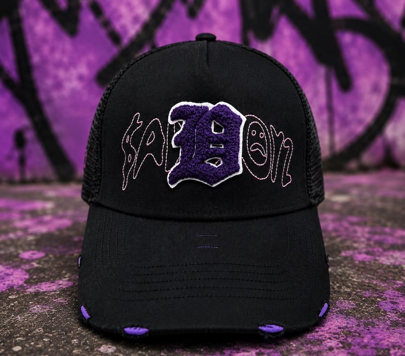 May include: A black trucker hat featuring a mesh back and a distressed brim with purple accents. The front panel displays a large, textured purple letter B patch with white trim, flanked by embroidered text that includes a sad face icon.