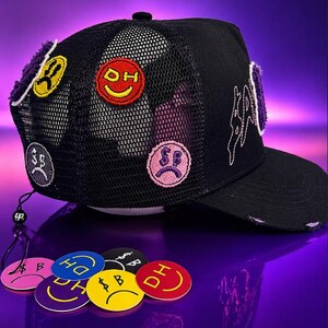 May include: A black trucker hat featuring a mesh back and multiple embroidered patches of colorful smiley faces with dollar signs and text. The hat has a curved brim with distressed edges and white embroidered text on the side panel.