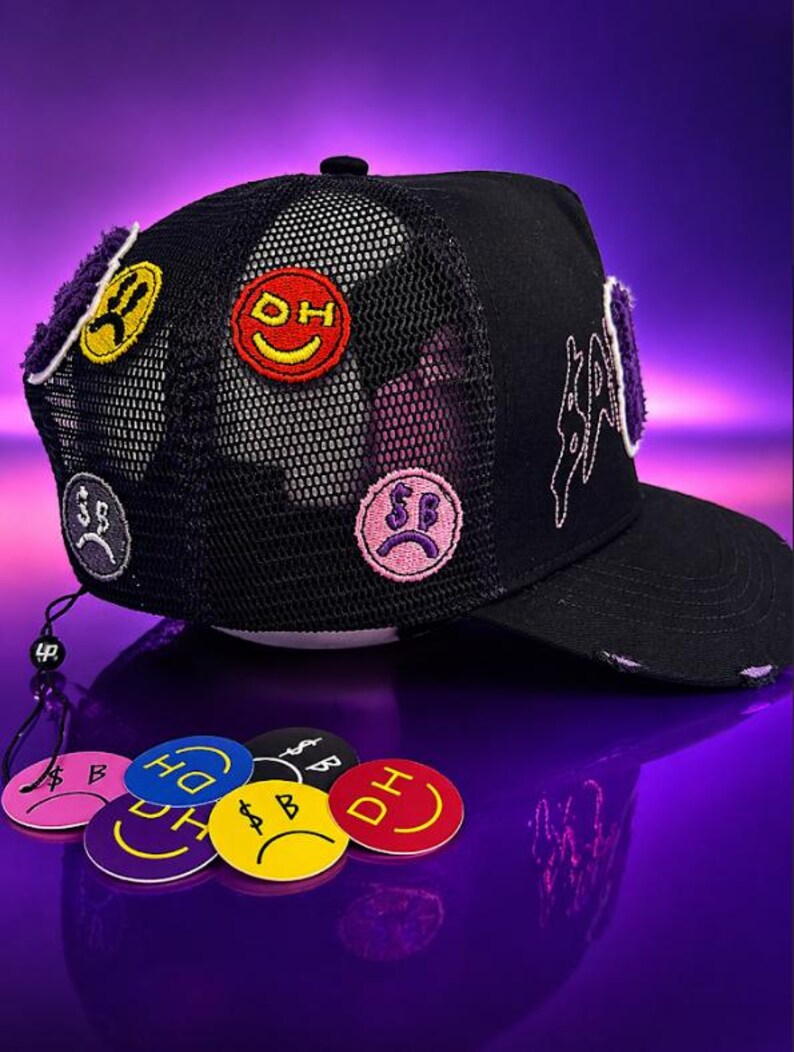 May include: A black trucker hat featuring a mesh back and multiple embroidered patches of colorful smiley faces with dollar signs and text. The hat has a curved brim with distressed edges and white embroidered text on the side panel.