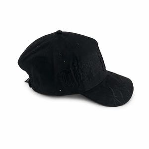 May include: A black baseball cap featuring tonal black embroidery on the front panel and a subtle flame-like pattern stitched onto the curved brim. The cap is constructed from a soft, matte material and includes a small black tag on the side.