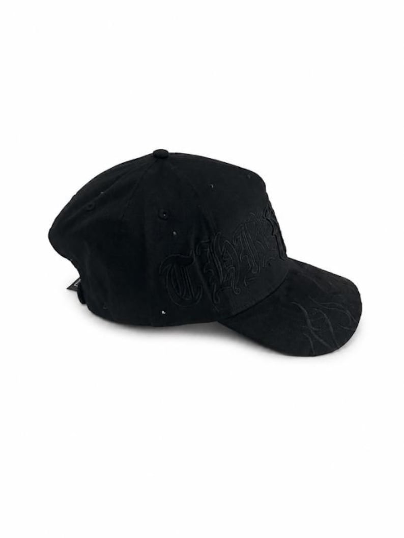 May include: A black baseball cap featuring tonal black embroidery on the front panel and a subtle flame-like pattern stitched onto the curved brim. The cap is constructed from a soft, matte material and includes a small black tag on the side.