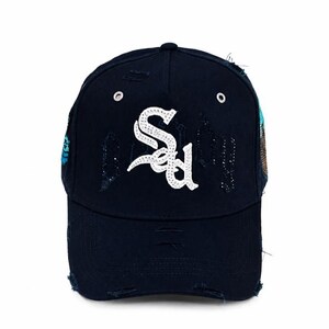 Könnte beinhalten: Eine dunkelmarineblaue Baseballkappe mit abgenutztem, ausgefranstem Finish und gezielten Rissen an Krone und Schirm. Die Vorderseite zeigt ein großes, weißes, mit Strasssteinen besetztes Monogramm-Logo sowie silberne Metallösen zur Belüftung.
