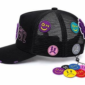 May include: A black trucker hat with a mesh back, featuring embroidered circular patches with smiley and sad faces, including text like DH and SB. The brim has distressed edges with purple fabric showing through, and several circular tags are attached.