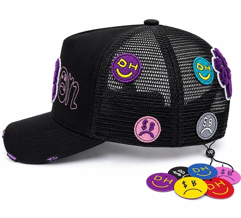 May include: A black trucker hat with a mesh back, featuring embroidered circular patches with smiley and sad faces, including text like DH and SB. The brim has distressed edges with purple fabric showing through, and several circular tags are attached.