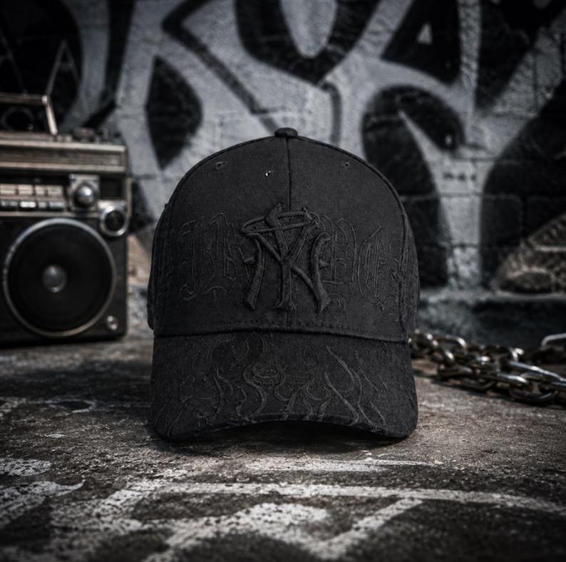 May include: A black baseball cap featuring a raised, embroidered logo on the front panel that resembles the letters NY. The cap includes tonal, subtle flame-like embroidery patterns across the crown and the curved brim, creating a monochromatic look.