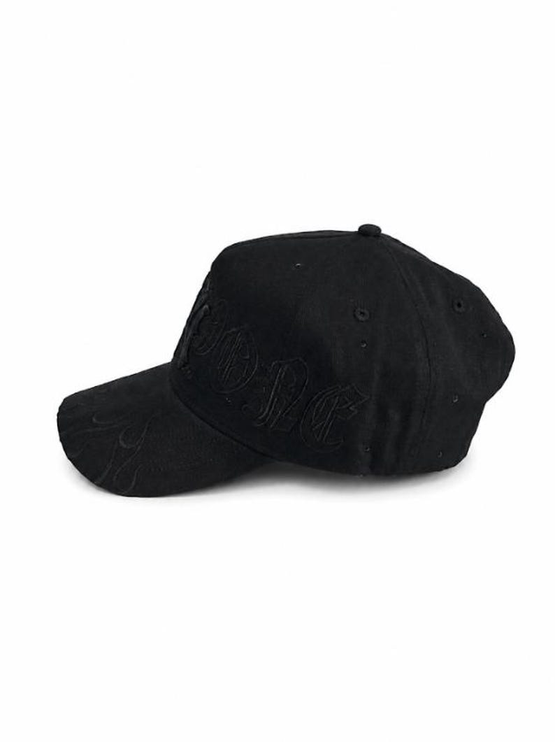 May include: A black baseball cap featuring tonal embroidery on the front panel and a subtle flame pattern stitched onto the curved brim. The cap has a structured crown with ventilation eyelets and a button at the top, all in a matching black fabric.