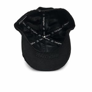 May include: A black baseball cap viewed from the inside, featuring a satin-like lining printed with the text ThirtyOne repeatedly. The word ThirtyOne is also embroidered in black on the underside of the brim, with a metal button at the crown.