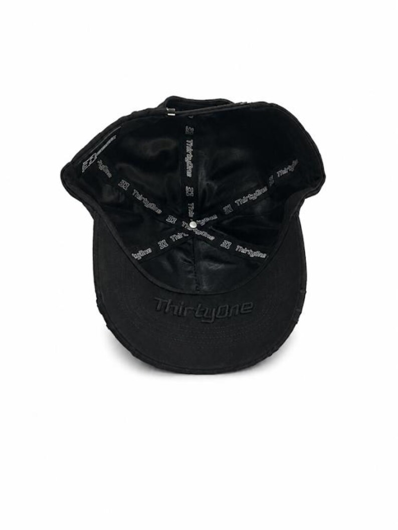 May include: A black baseball cap viewed from the inside, featuring a satin-like lining printed with the text ThirtyOne repeatedly. The word ThirtyOne is also embroidered in black on the underside of the brim, with a metal button at the crown.