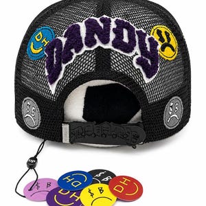 May include: A black mesh trucker hat featuring the word DANDY in purple fuzzy lettering across the front. The hat is decorated with circular patches showing smiley and sad faces. A black plastic snapback strap and several loose circular tags are included.