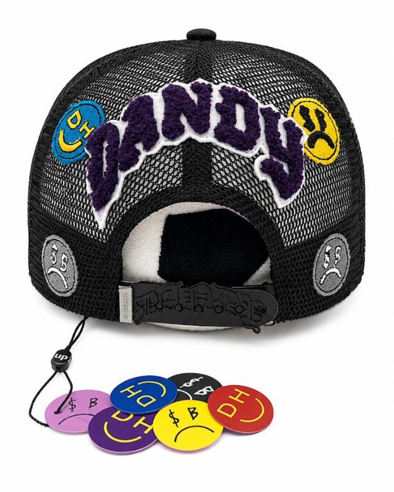 May include: A black mesh trucker hat featuring the word DANDY in purple fuzzy lettering across the front. The hat is decorated with circular patches showing smiley and sad faces. A black plastic snapback strap and several loose circular tags are included.