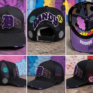 May include: A black trucker hat featuring purple embroidered text that reads "BADBOY" and "DANDY," accented with colorful circular patches depicting various facial expressions. The hat includes a mesh back, a distressed brim, and interior lining.