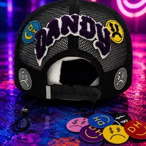May include: A black mesh trucker hat featuring the word DANDY in large purple chenille letters across the front. The hat is decorated with various circular patches displaying smiley faces, sad faces, and the letters DH or dollar signs in multiple colors.