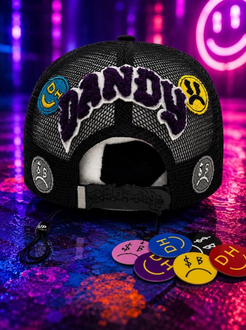 May include: A black mesh trucker hat featuring the word DANDY in large purple chenille letters across the front. The hat is decorated with various circular patches displaying smiley faces, sad faces, and the letters DH or dollar signs in multiple colors.