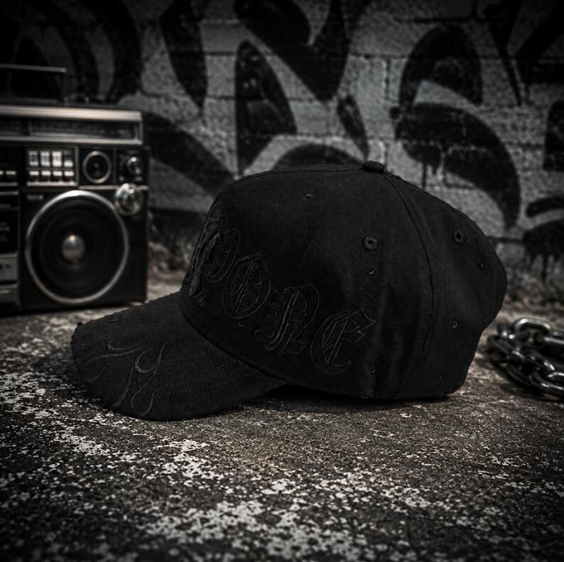 May include: A black baseball cap featuring tonal embroidery on the front panel with the word "RIPONE" in a gothic-style font. The curved brim displays subtle, matching black flame-pattern stitching, and the cap includes ventilation eyelets.