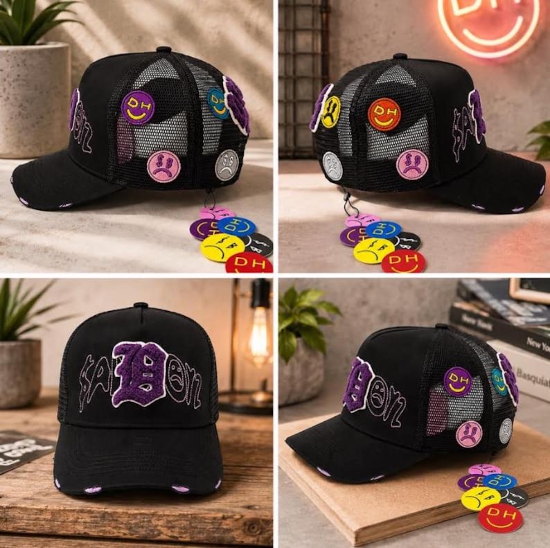 May include: A black trucker hat featuring a mesh back and a curved brim. The front displays purple embroidered text reading "SAIBON" with a sad face icon. The sides are adorned with various colorful circular patches and dangling charms.