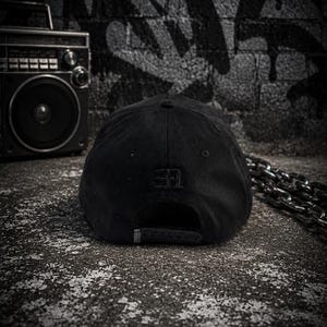 May include: A solid black baseball cap featuring a structured crown and a small, tonal embroidered logo on the back panel. The cap includes a metal buckle closure at the rear, providing a clean, monochromatic look with subtle stitching details throughout.
