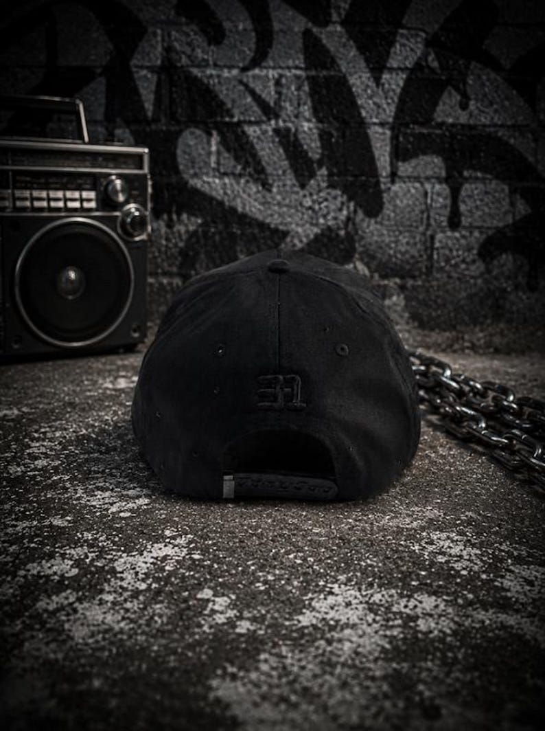 May include: A solid black baseball cap featuring a structured crown and a small, tonal embroidered logo on the back panel. The cap includes a metal buckle closure at the rear, providing a clean, monochromatic look with subtle stitching details throughout.