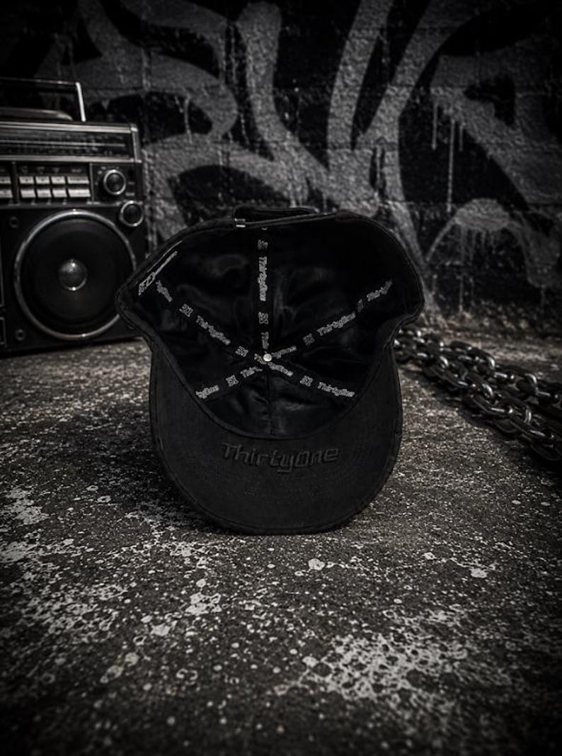 May include: A black baseball cap shown from the interior, featuring a smooth satin-like lining with the text ThirtyOne printed repeatedly on the inner seam tape. The word ThirtyOne is also embroidered in black thread on the underside of the brim.