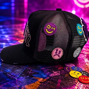May include: A black trucker hat featuring a mesh back panel and a solid front panel. The hat is decorated with several circular embroidered patches depicting various smiley and sad faces in purple, pink, blue, and gray, along with the letters DH.