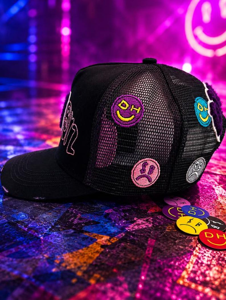 May include: A black trucker hat featuring a mesh back panel and a solid front panel. The hat is decorated with several circular embroidered patches depicting various smiley and sad faces in purple, pink, blue, and gray, along with the letters DH.