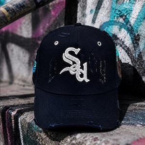 May include: A black baseball cap featuring a distressed, worn-in finish with frayed edges. The front panel displays the letters "SD" in a large, stylized font encrusted with clear rhinestones. The cap includes metal eyelets for ventilation.