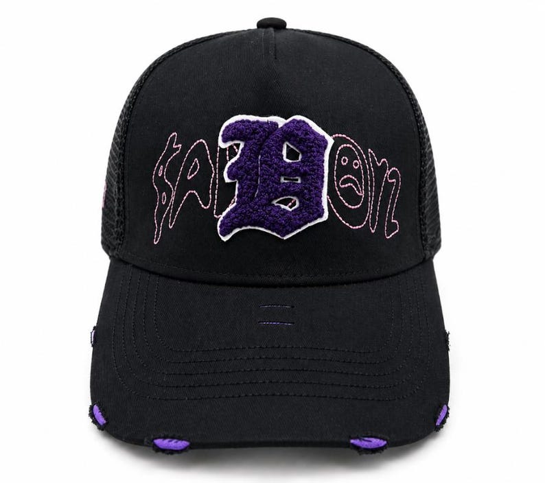May include: A black trucker hat featuring a mesh back and a structured front panel. The front displays the text "SADBOYZ" in pink stitched lettering with a large, textured purple chenille letter "B" in the center. The brim has distressed edges with purple fabric accents.