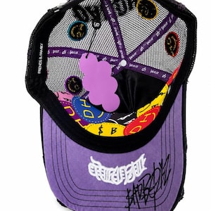 May include: A purple and black trucker hat with a mesh back, featuring a purple cloud-shaped hang tag. The interior lining displays colorful circular patches with text, including "FRIENDS & FAMILY" on the inner band and white embroidered graffiti-style text on the front brim.