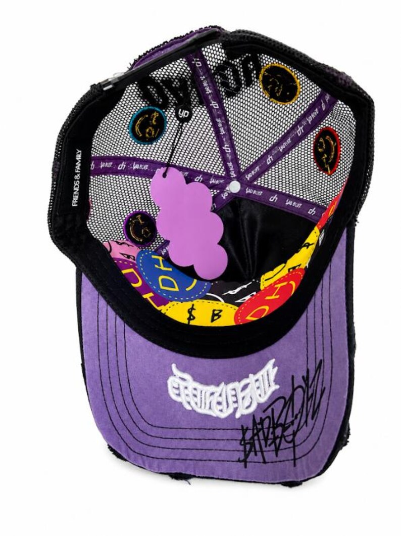 May include: A purple and black trucker hat with a mesh back, featuring a purple cloud-shaped hang tag. The interior lining displays colorful circular patches with text, including "FRIENDS & FAMILY" on the inner band and white embroidered graffiti-style text on the front brim.